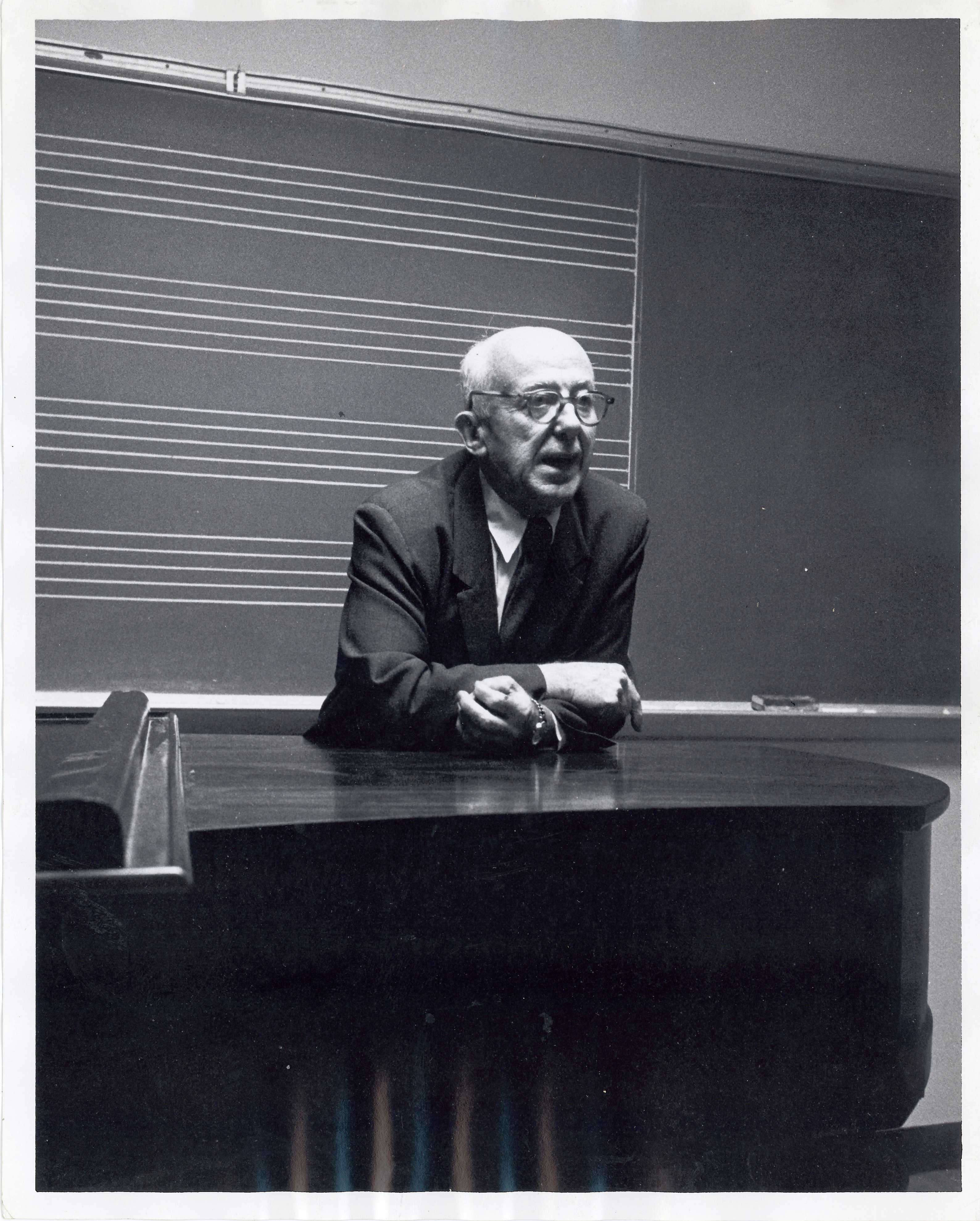 A man leaning on a baby grand piano in front of a blackboard.