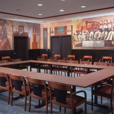 Room with a table surrounded by chairs and murals on the walls. 