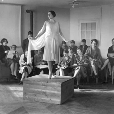 A black and white photo of a fashion studies class viewing a model in a white dress on a platform. 